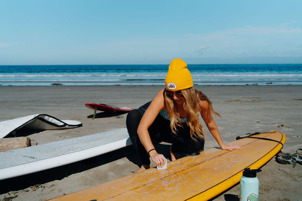 girl waxes surfboard while wearing yellow beanie with alki patch
