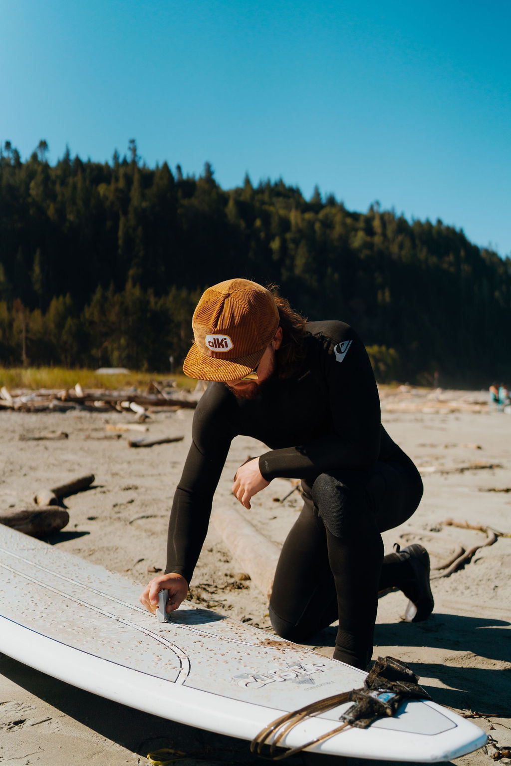 surfer in wetsuit wears brown snapback corduroy hat while waxing surfboard